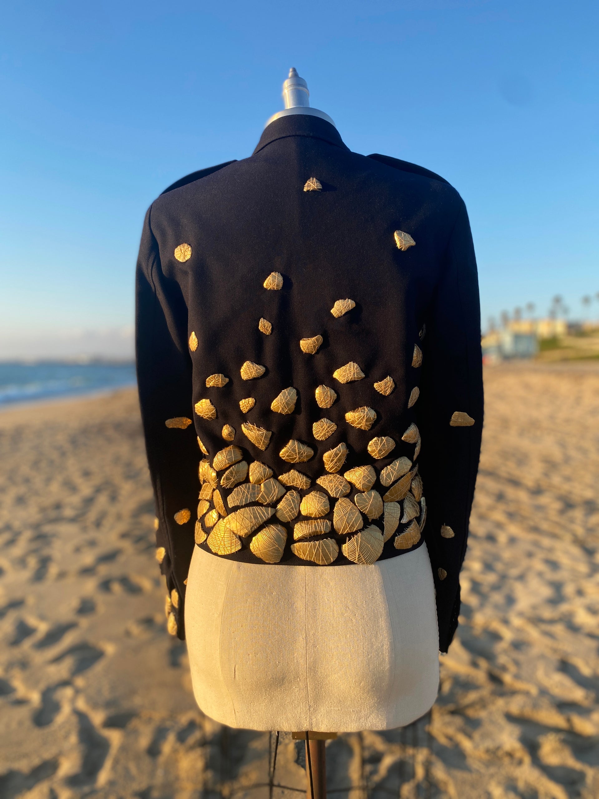 Back view of Shireen Renée's 'Shell Shock' Couture French Air Force Jacket, a cropped Navy jacket adorned with numerous gold shells, is displayed on a mannequin. The mannequin stands on a sandy beach, with the ocean and a clear blue sky visible in the background, creating a striking contrast between the textured jacket and the natural setting.