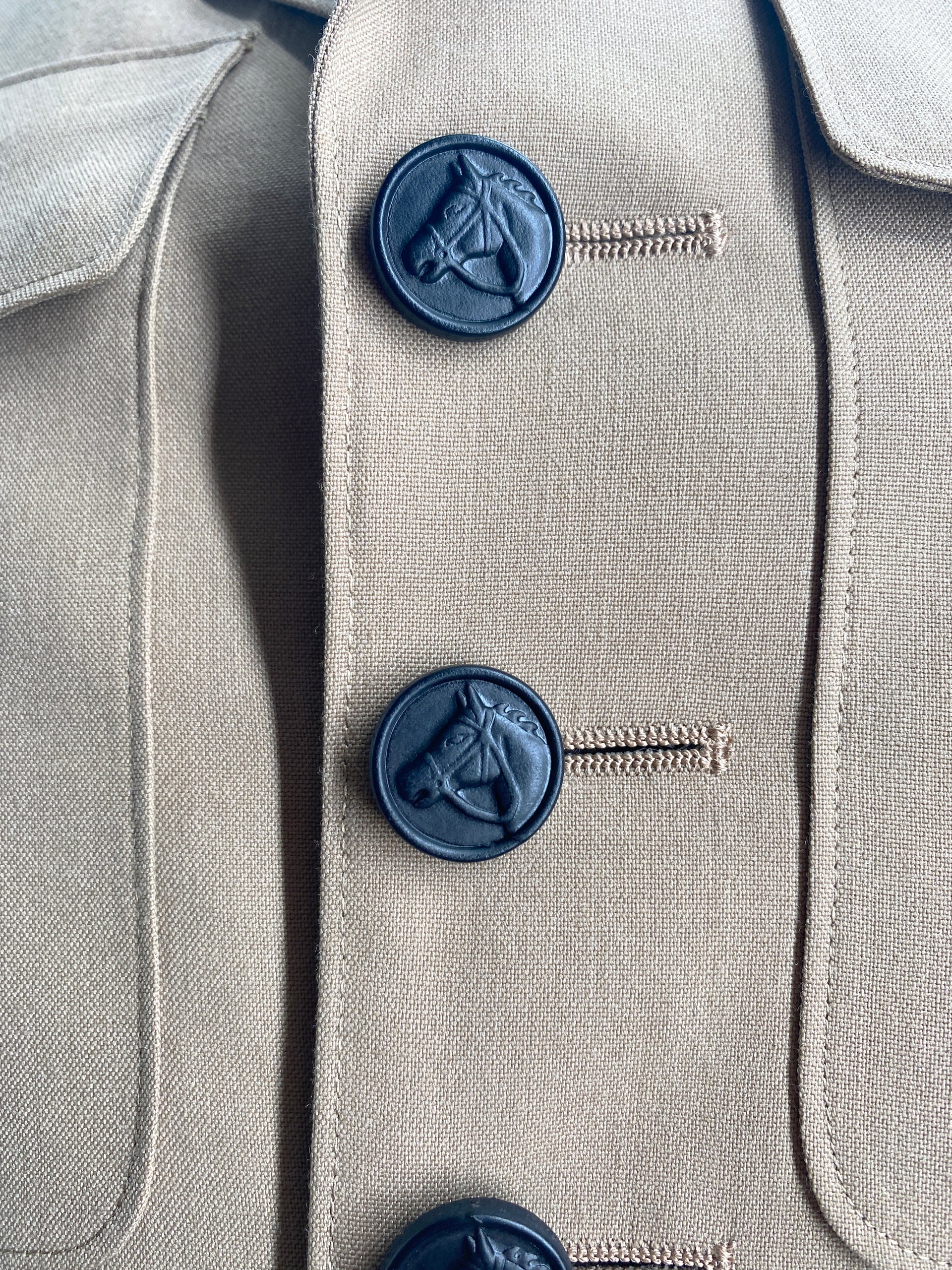 A textured close-up of a jacket, highlights three round, matte black buttons. Each button features a detailed, raised design of a horse's head in profile. The buttons are vertically aligned, with visible stitching securing them to the fabric. The overall image emphasizes the texture and craftsmanship of the garment.