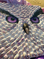 Textured owl face art by Shireen Renee, mixed media art using pistachio shells, with purple eyes, resting on sand with a draped purple patterned fabric. Unique animal artwork, beach setting, artistic composition.