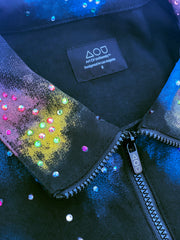 A close-up studio shot of a dark blue, collared jacket with a full zipper. The jacket features a vibrant, abstract pattern of colorful splotches and dots in blue, purple, yellow, and white, creating a galaxy-like effect. The jacket is presented against a digital backdrop of space, with two planets visible in the background. This is an Art Of Uniformity brand jacket by Shireen Renee.
