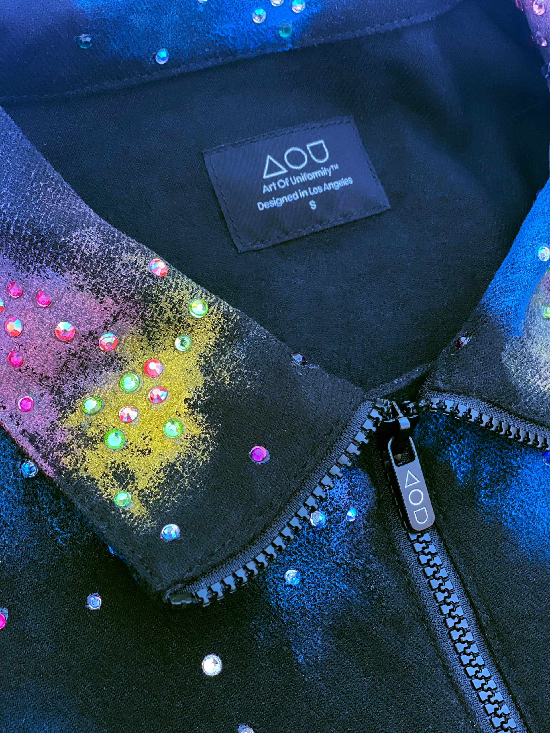 A close-up studio shot of a dark blue, collared jacket with a full zipper. The jacket features a vibrant, abstract pattern of colorful splotches and dots in blue, purple, yellow, and white, creating a galaxy-like effect. The jacket is presented against a digital backdrop of space, with two planets visible in the background. This is an Art Of Uniformity brand jacket by Shireen Renee.