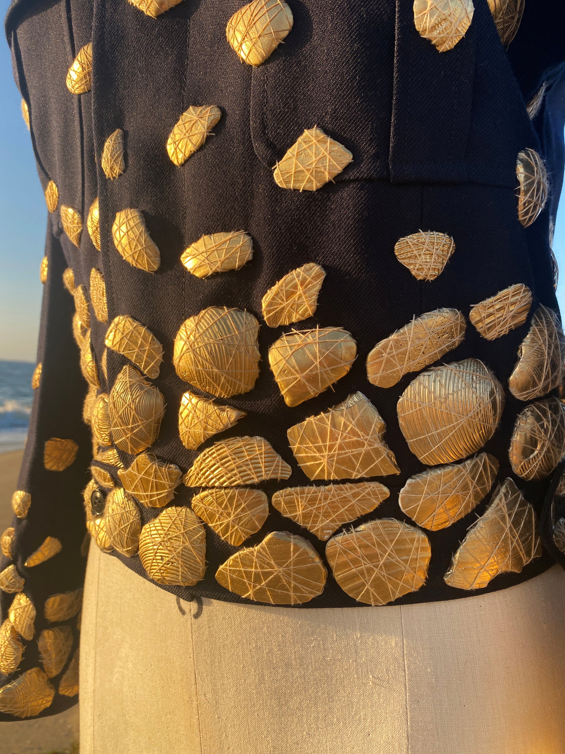 A close-up shot captures the texture and detail of a Navy jacket adorned with numerous gold shells. The shells are meticulously attached with delicate gold thread, forming intricate patterns. In the background, a glimpse of the ocean provides a natural contrast to the crafted garment.