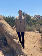 Front image of a model standing in a desert environment wearing a vintage American USMC khaki jacket with hand-embroidery line art on the back and gold-tipped tassels at the back vent. There are rock formations in the distance.