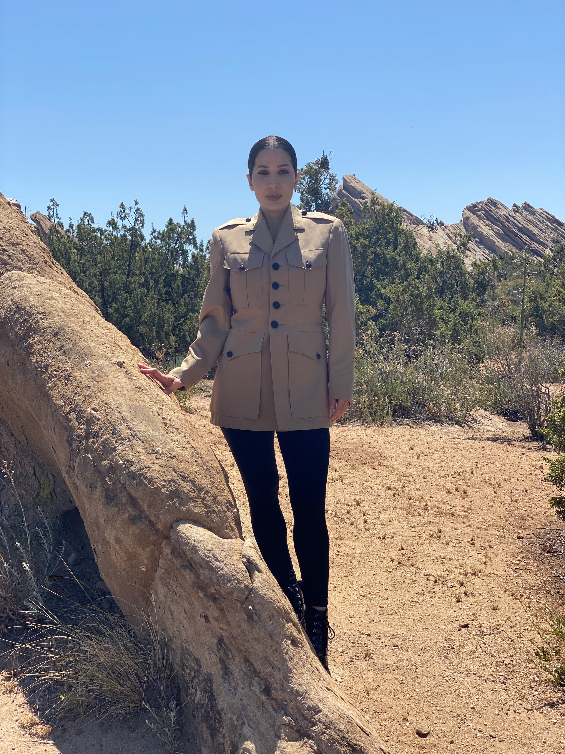 Front image of a model standing in a desert environment wearing a vintage American USMC khaki jacket with hand-embroidery line art on the back and gold-tipped tassels at the back vent. There are rock formations in the distance.