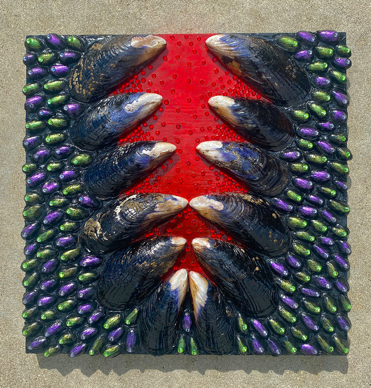 Close-up of a mixed media artwork by Shireen Renee, showing mussel shells, painted pistachio shells in purple and green, and rhinestones arranged on a wood panel with a red stripe down the center.