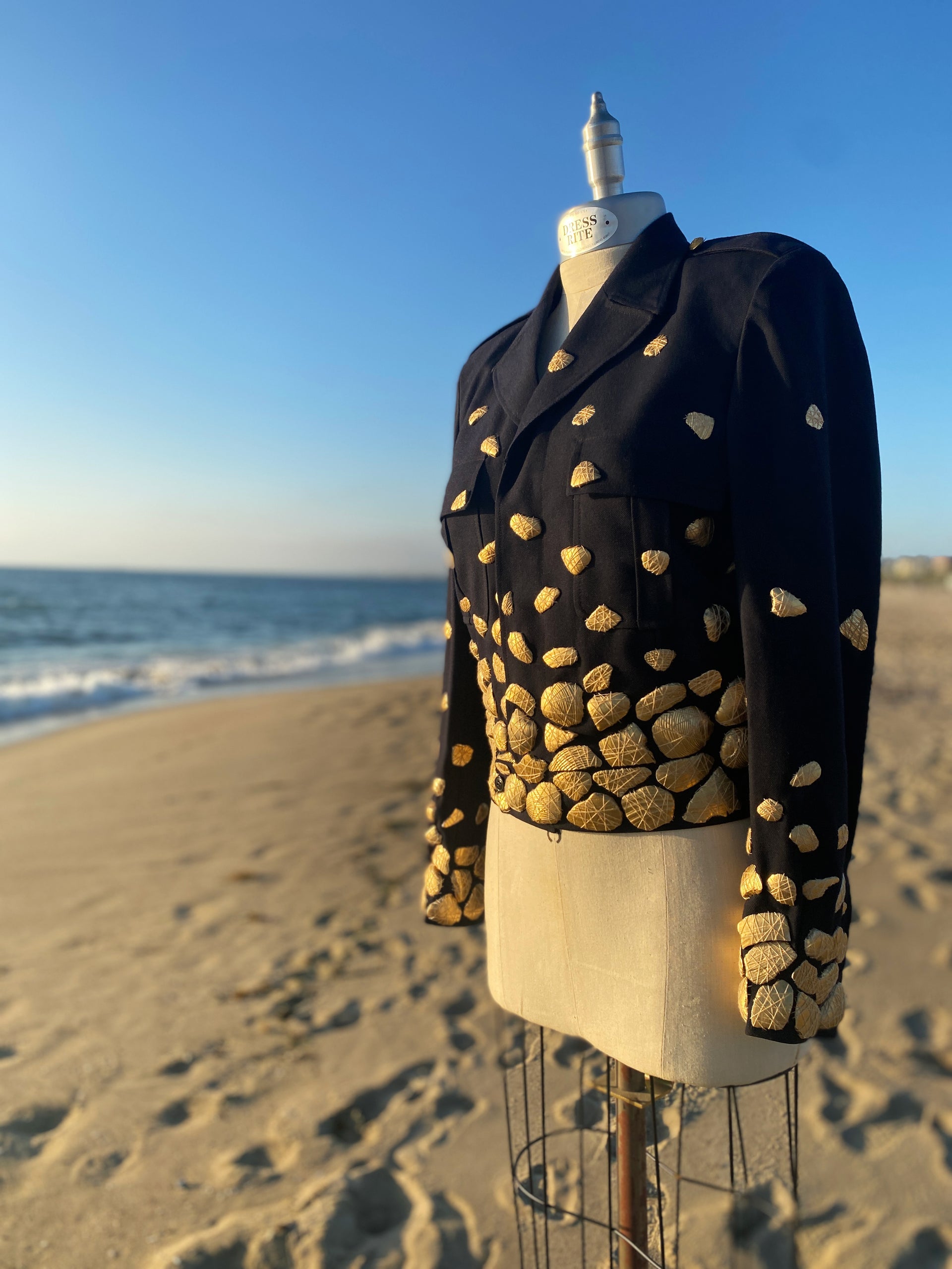Shireen Renée's 'Shell Shock' Couture French Air Force Jacket, a cropped Navy jacket adorned with numerous gold shells, is displayed on a mannequin. The mannequin stands on a sandy beach, with the ocean and a clear blue sky visible in the background, creating a striking contrast between the textured jacket and the natural setting.