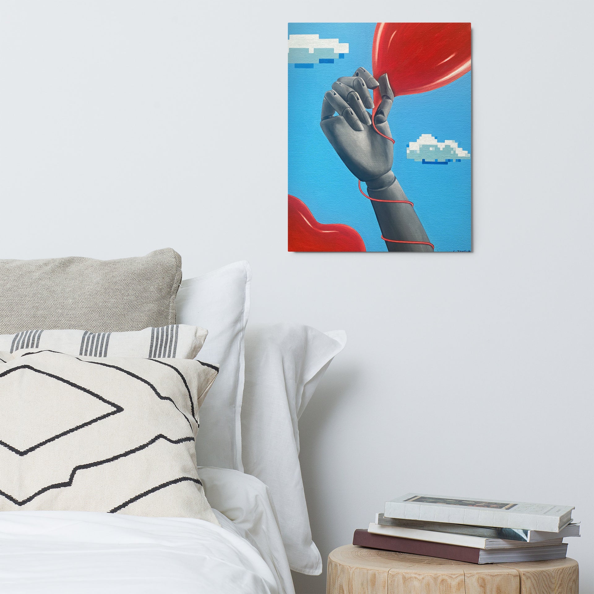 Fine art contemporary metal print by Shireen Renee of a mannequin hand holding a heart-shaped balloon against a sky blue background with pixelated clouds. The artwork is hanging on a plain white wall in a bedroom to the right of a bed with white linens and above a wooden table with a stack of books.