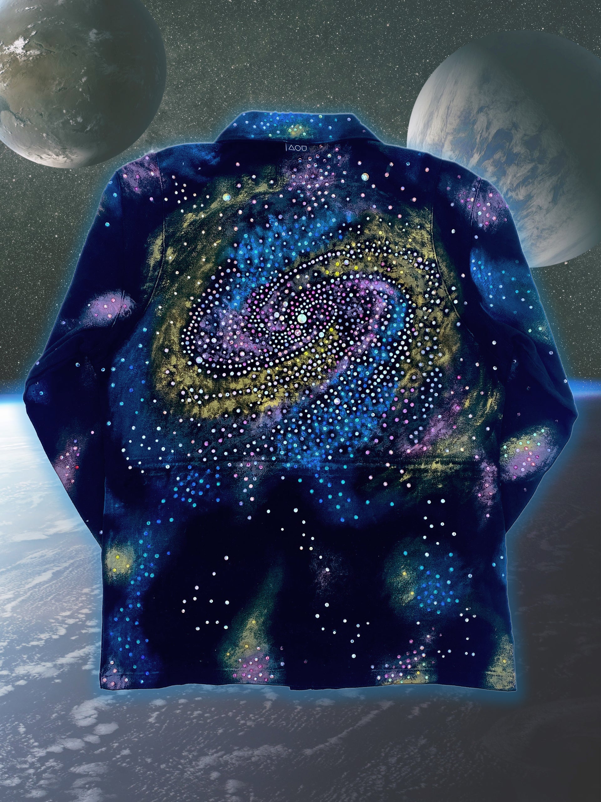 A close-up studio shot of a dark blue, collared jacket with a full zipper. The jacket features a vibrant, abstract pattern of colorful splotches and dots in blue, purple, yellow, and white, creating a galaxy-like effect. The jacket is presented against a digital backdrop of space, with two planets visible in the background. This is an Art Of Uniformity brand jacket by Shireen Renee.