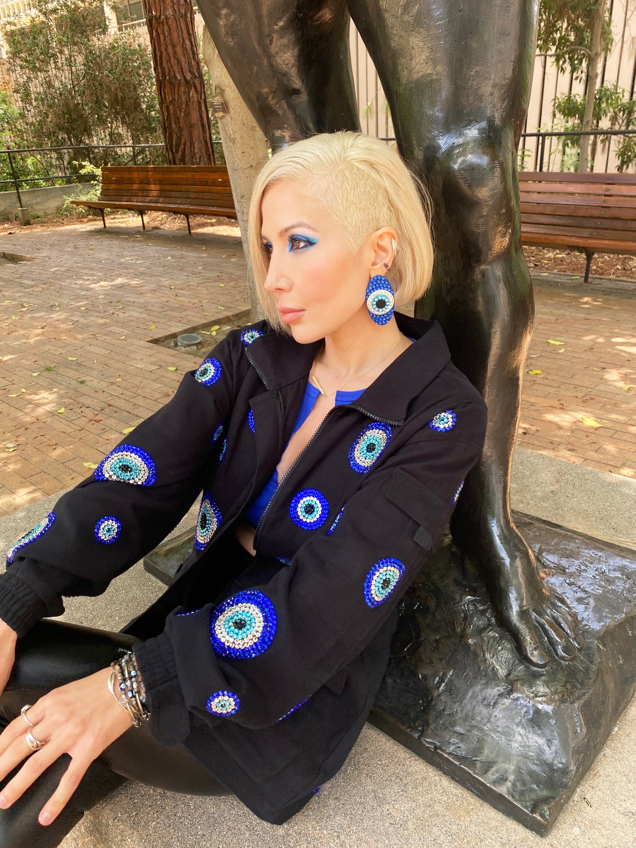 A woman with short blonde hair (shaved on the right side) and striking blue eye makeup poses next to a dark bronze statue, which shows the base of its legs. She is wearing a black jacket adorned with numerous round evil eye designs in various shades of blue, white, and black, accented with rhinestones. 