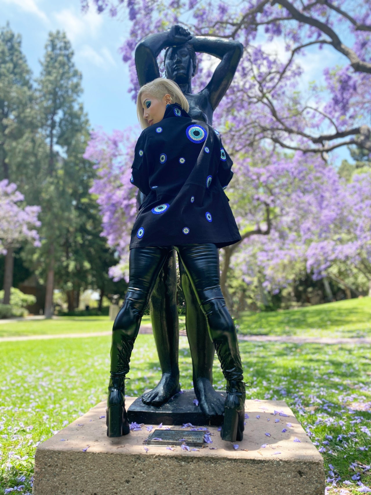 A woman with short blonde hair and bright blue eye makeup is shown from behind a dark bronze statue. She is wearing a black jacket adorned with numerous blue and white rhinestone evil eye designs, and black shiny pants and boots. Their head is turned to the left, looking away from the camera. The setting is outdoors with a green grassy area and trees with purple flowers in bloom in the background.