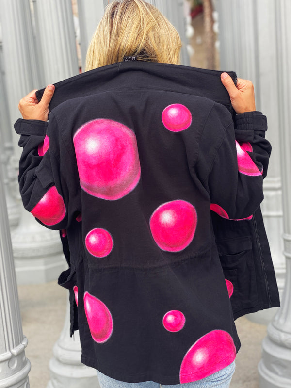 Back view of blonde model wearing the Art Of Uniformity "Pink Dreams" black organic jacket. The Jacket is black with giant pink hand-painted bubbles all over.