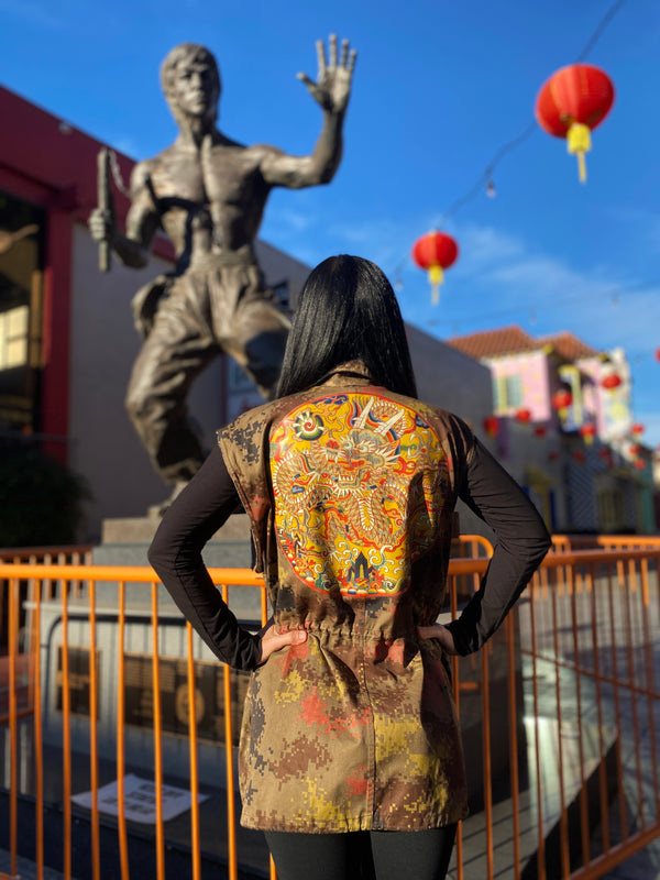 Model standing in front of a Bruce Lee statue in DTLA Chinatown wearing a Chinese digital camo jacket with a large dragon medallion art print on the back.
