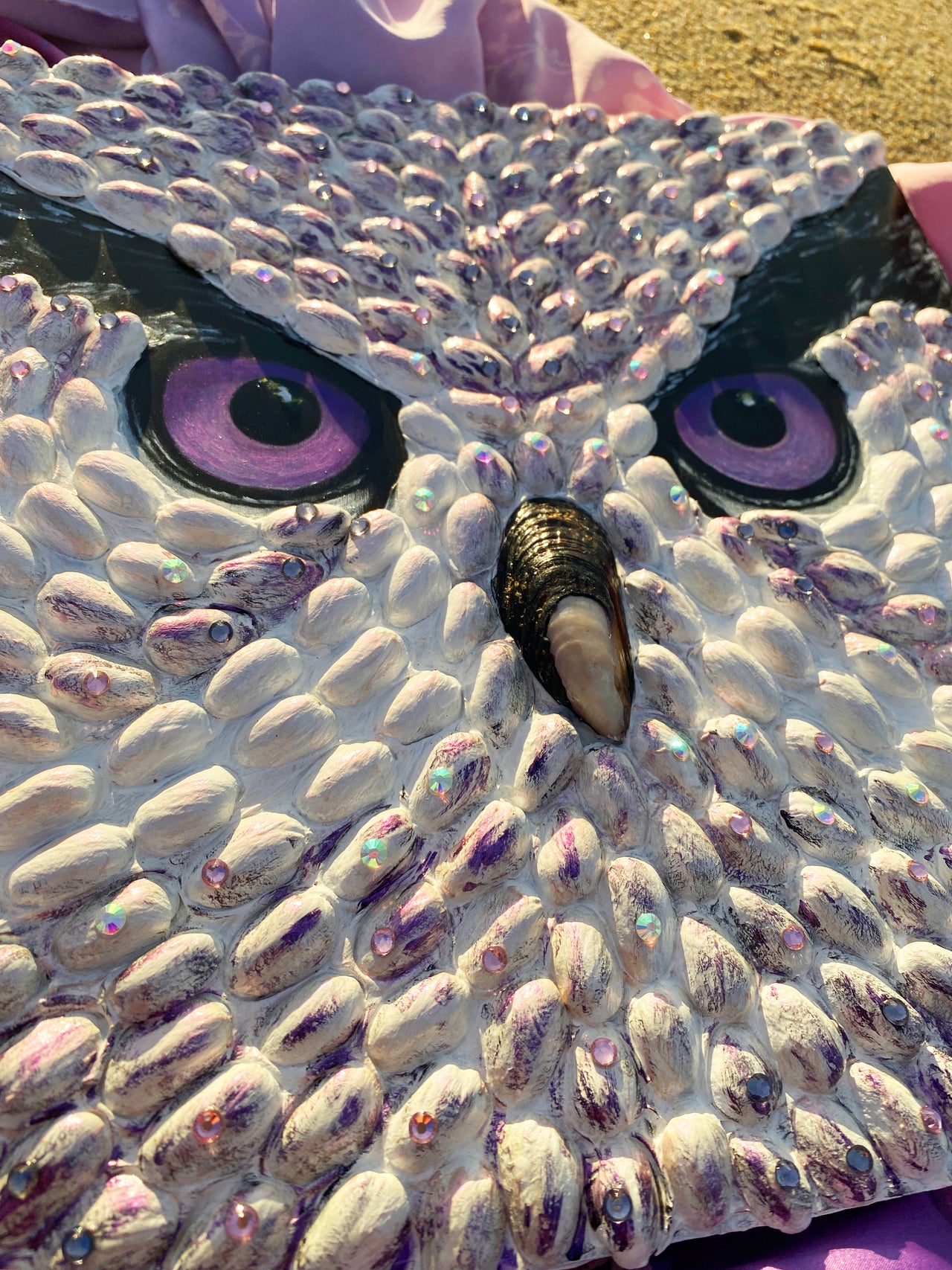 Textured owl face art by Shireen Renee, mixed media art using pistachio shells, with purple eyes, resting on sand with a draped purple patterned fabric. Unique animal artwork, beach setting, artistic composition.