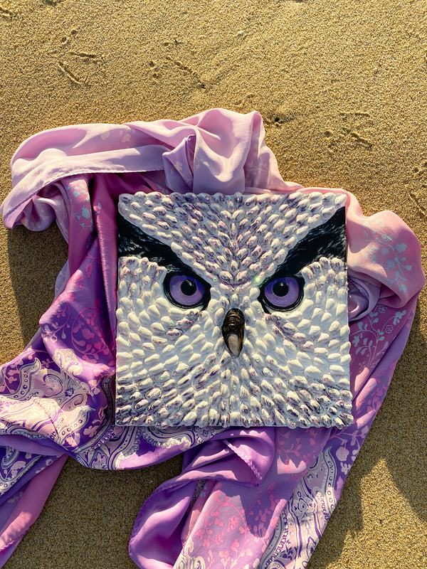 Textured owl face art by Shireen Renee, mixed media art using pistachio shells, with purple eyes, resting on sand with a draped purple patterned fabric. Unique animal artwork, beach setting, artistic composition.