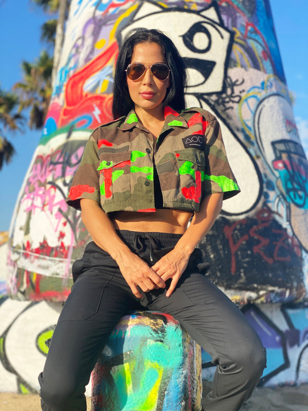 Model sitting in front of a graffiti structure wearing a vintage French cropped camo button-up shirt hand-painted with neon orange and green paint.