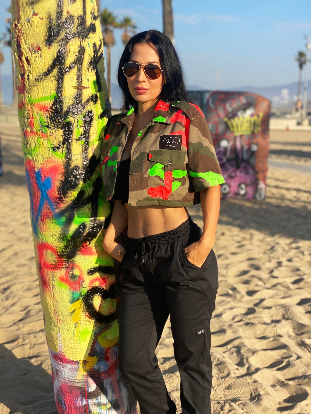 Model standing next to a graffitied palm tree in Venice Beach, CA wearing a vintage French cropped camo button-up shirt hand-painted with neon orange and green paint.