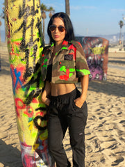 Model standing next to a graffitied palm tree in Venice Beach, CA wearing a vintage French cropped camo button-up shirt hand-painted with neon orange and green paint.