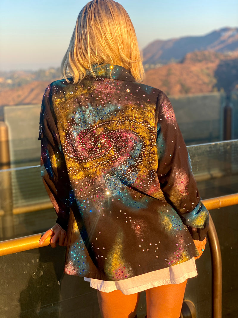 An image of a blond woman standing next to a ledge at the Griffith Observatory. Her back is to the viewer and she is wearing a black, collared jacket with a full zipper. The jacket features a vibrant, abstract pattern of colorful splotches and dots in blue, purple, yellow, and white, creating a galaxy-like effect. The jacket is presented against a digital backdrop of space, with two planets visible in the background. This is an Art Of Uniformity brand jacket by Shireen Renee. 