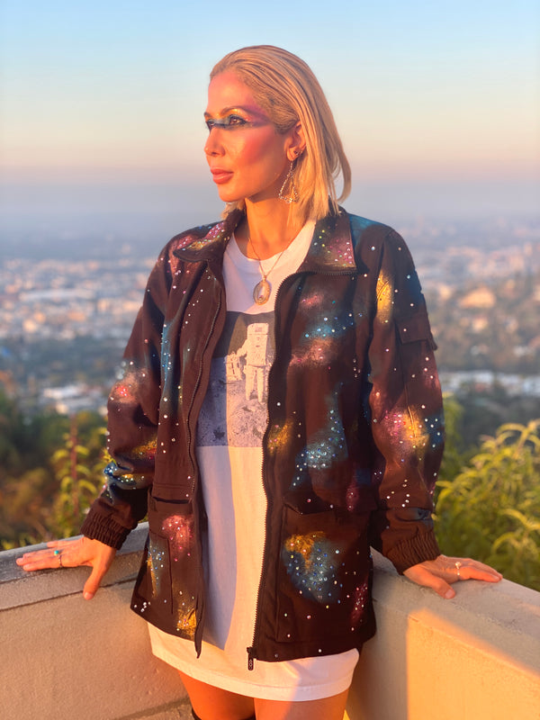 An image of a blond woman standing next to a ledge with the city scape of Los Angeles behind her. She is wearing a black, collared jacket with a full zipper. The jacket features a vibrant, abstract pattern of colorful splotches and dots in blue, purple, yellow, and white, creating a galaxy-like effect. The jacket is presented against a digital backdrop of space, with two planets visible in the background. This is an Art Of Uniformity brand jacket by Shireen Renee. 