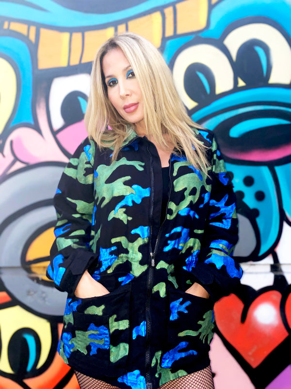 Model standing in front of a colorful mural wearing the Art Of Uniformity "Camo Planet" organic black brushed cotton unisex jacket with blue and green hand-painted camo print.