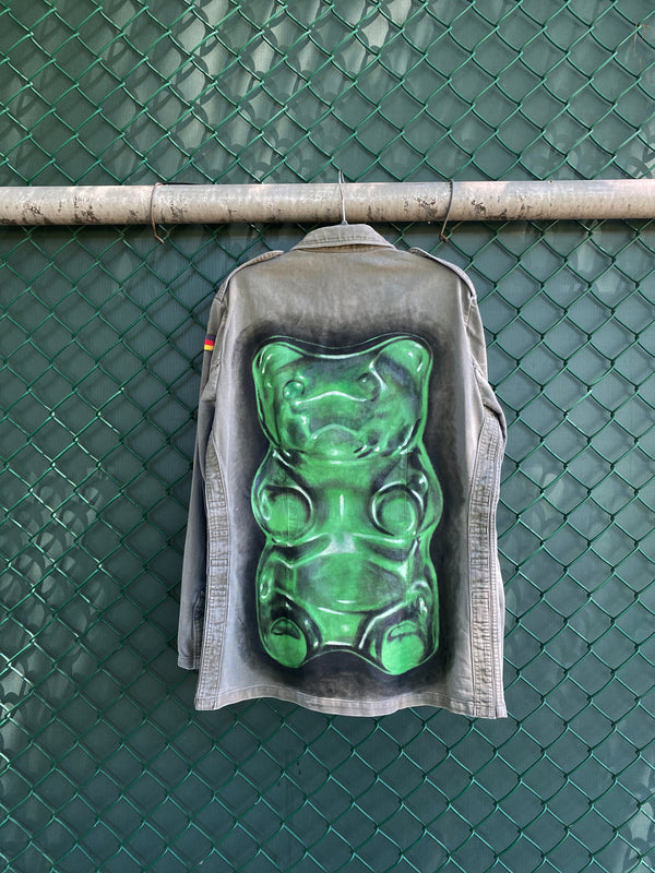 Artwear gummy bear jacket hanging on a chain link fence with back of jacket showing. Jacket showcases a giant, painted green gummy bear by Los Angeles artist, Shireen Renee.